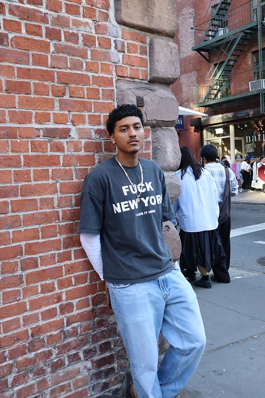 Graphic tee and light denim, leaning on brick.